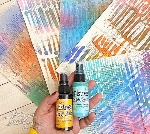 A hand holding two bottles of spray ink and surrounded by colorful sprayed papers.