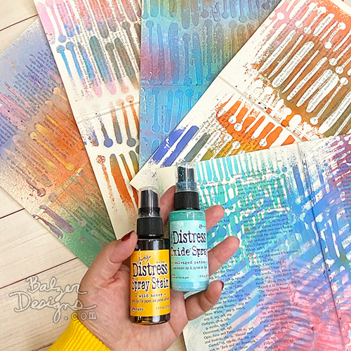 A hand holding two bottles of spray ink and surrounded by colorful sprayed papers.