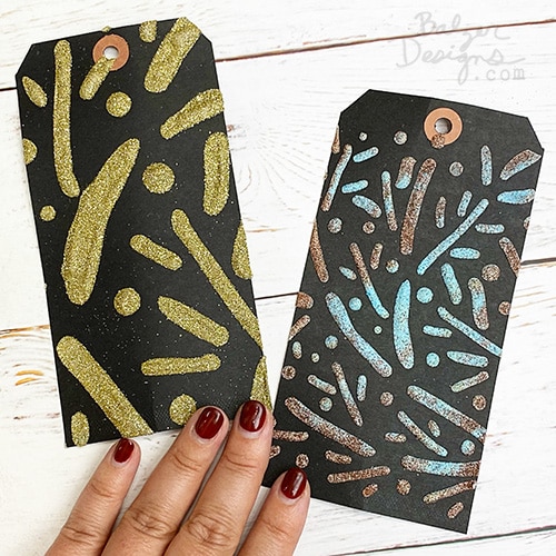 Two black tags with a stenciled design that has been heat embossed. There are four fingers (with red nails) laying on the tags.